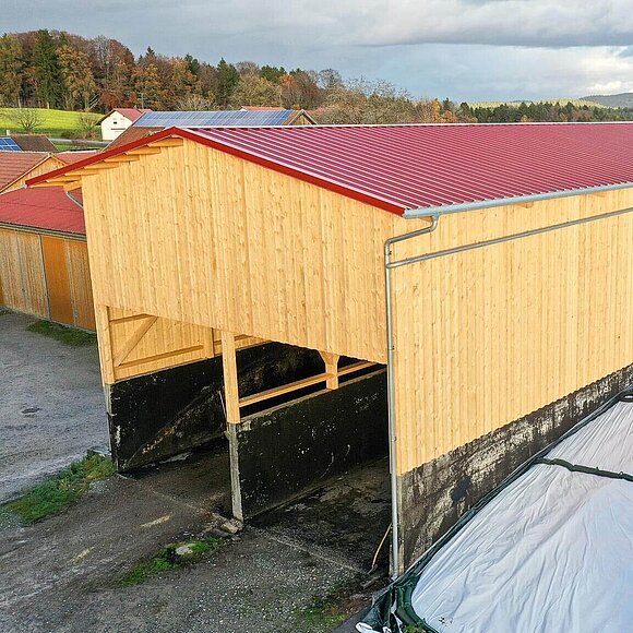 Langer Holzbau mit rotem Dach in ländlicher Umgebung, teilweise fertiggestellt.